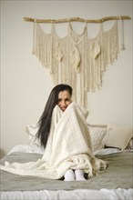 Smiling woman sitting on the bed, wrapped in a soft blanket