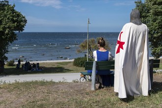 A knight with a red cross on a white coat and armour stands behind a woman sitting on a bench with