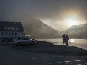 Sunrise on a foggy morning, hotel Grimsel mountain pass at lake Totensee, couple taking pictures,