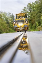 Yellow track construction train on rails, reflected in the water, surrounded by trees, track