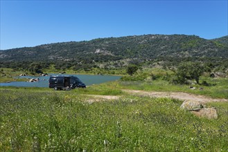 Motorhome parked on a green meadow near a lake, surrounded by mountains and trees on a sunny day,