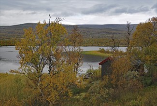 A hut stands on the shore of a lake, surrounded by autumnal trees and mountains in the background,