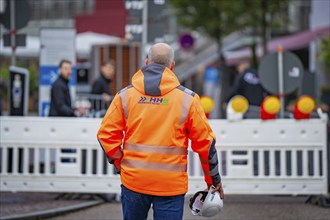 A worker in an orange safety waistcoat with a helmet stands in front of a barrier on the road,