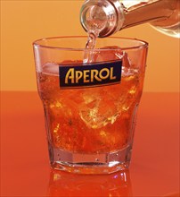 Aperol on ice cube cubes in an aperitif tumbler against an orange background is topped up with soda