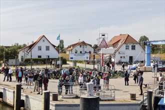 People gather at the harbour next to buildings and bicycles, a flag in the background, Rügen,