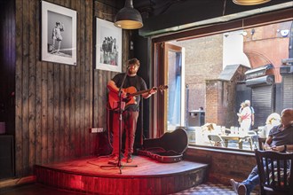 A guitarist performs in a cosy bar with wooden walls and large windows, Liverpool