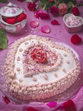 A heart-shaped cake decorated in pink and white, surrounded by petals, roses, flying sugar