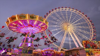 A lively funfair at dusk, illuminated by a large Ferris wheel and a colourful chain carousel,