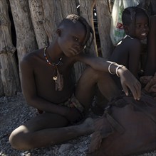 Himba girl with braided pigtails sitting in the shade near a hut, near Opuwo, Kaokoveld, Kunene,