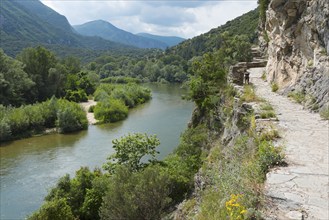 A rocky hiking trail along the bank of a river with surrounding mountain views, River Nestos or