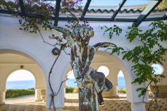 Bronze angel statue in front of arched windows overlooking the sea, decorated with plants and round