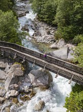 A hiker crosses a wooden bridge in a forest with a raging river flowing underneath, Zillertal,