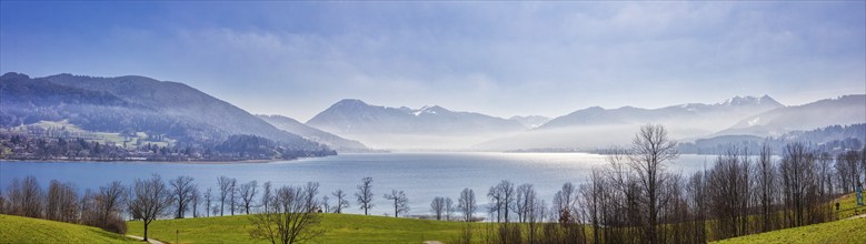 A picturesque lake surrounded by mountains and trees under a blue, slightly misty sky, Tegernsee in