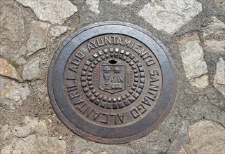 A round metal manhole cover with a coat of arms and text on a paved surface, manhole cover, gully