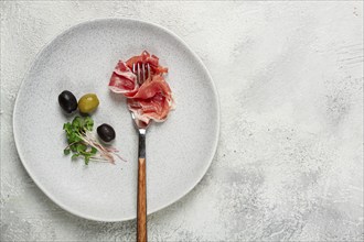 Prosciutto on a fork, top view, olives and micro-greens on a plate, light background, no people