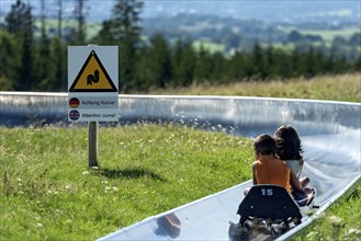 Boy and girl with sledges on the summer toboggan run, warning sign Attention curve, ski and