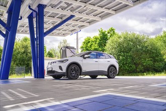 An electric car at a charging station under a modern roof with solar panels, surrounded by green