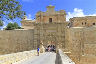 Tourists walking over moat bridge through entrance gateway of medieval city of Mdina, Malta, Europe