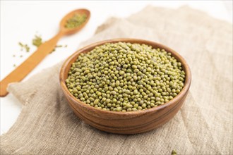 Wooden bowl with raw green mung bean and wooden spoon on a white background and linen textile. Side