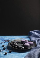 Purple macarons or macaroons cakes with blueberries on ceramic plate on a blue and black background