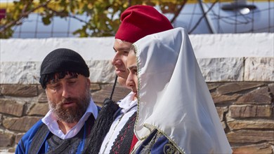 Three people in traditional historical Cretan dress stand next to each other in conversation, visit