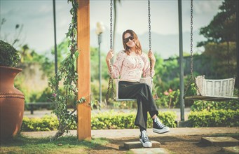 Happy young woman sitting on a swing in a nature park. Portrait of a smiling girl sitting on a