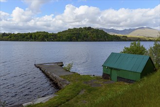 A scenic lake view with a green shed beside a dock, surrounded by hills, trees, and a partly cloudy