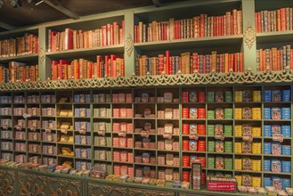 Wall of shelves with books on top and numerous colourful tins underneath, tins of sardines, tinned