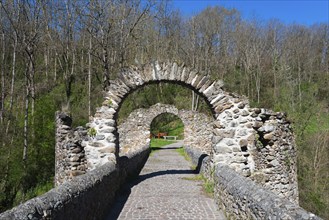 Steinbrücke mit alten Mauern und Torbogen inmitten eines grünen Waldes, unter blauem Himmel, Brücke