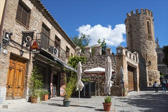 Medieval stone building with tower, shops, parasols and plants, on a street under a blue sky,