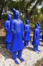 A group of blue terracotta warrior statues stands on gravel, surrounded by trees, Bacalhôa,