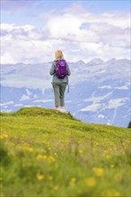 Person with rucksack looking into a wide mountain landscape, standing on a green meadow, Penken,