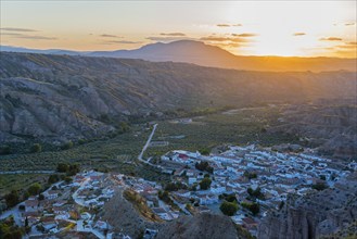 A village lies in a valley surrounded by hills and mountains as the sun sets and the valley glows