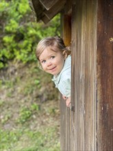 A cheerful child looks out of a wooden house in a green forest setting