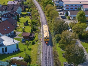 Yellow train runs through village, surrounded by houses, gardens and trees, tamping machine,