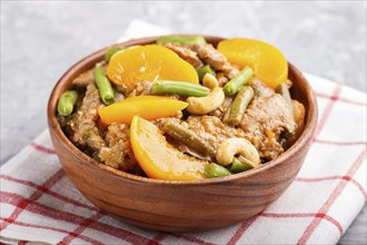 Fried pork with peaches, cashew and green beans in a wooden bowl on a gray concrete background.