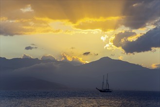 A sailboat on the sea in front of a dramatic sunset, yellow rays of light over the mountains, Agia