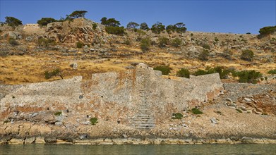 Old stone wall on rocky cliff with dry vegetation and calm sea water in the foreground, Venetian