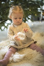 A toddler holds a glittering Christmas bauble in front of a Christmas tree in a cosy, wintry