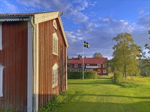 Red wooden house on a green meadow with a flag next to it, under a blue sky with clouds, STF home