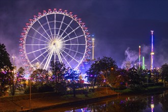 An illuminated Ferris wheel and fairground rides at night, surrounded by trees by the water,