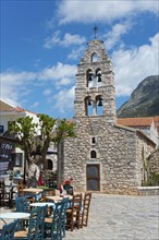 Traditional stone church with bell tower next to a café with terrace under a clear blue sky