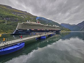 A large cruise ship, Mein Schiff 6, lies in the fjord, surrounded by green mountains under a cloudy