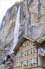 An impressive waterfall cascades down next to a wooden house with green shutters. A steep rock face