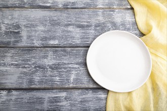 Empty white ceramic plate on gray wooden background and yellow linen textile. Top view, copy space,