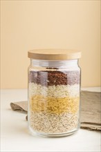 Glass jar with different kinds of rice poured in layers on white and orange background. side view,