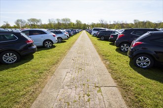 A wide paved walkway stretches between rows of parked cars in an outdoor lot with green grass