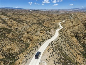 A lonely road leads through barren, hilly desert terrain with a steep rocky backdrop and blue sky,