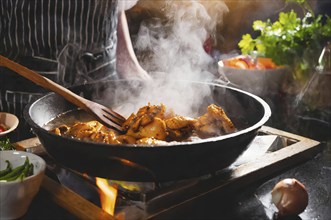 A person is cooking chicken in a pan on a stove. The pan is filled with a red sauce and the chicken