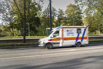 An ambulance drives fast along a road surrounded by autumnal trees, Stuttgart, Germany, Europe